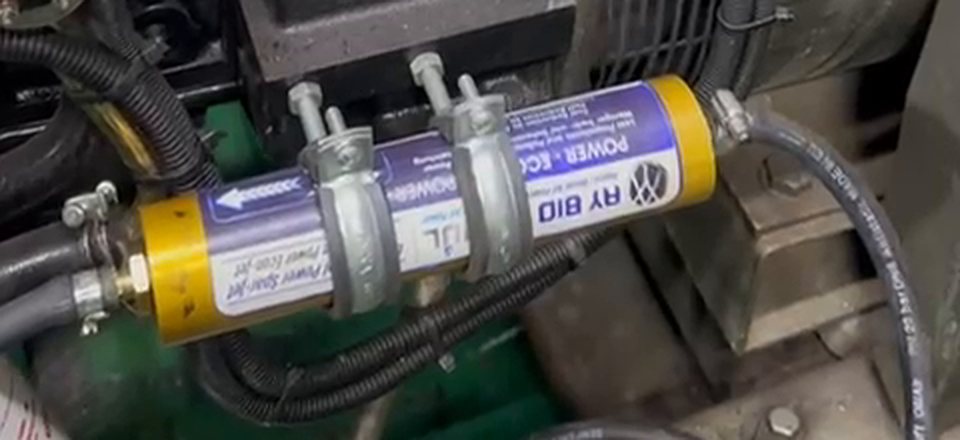 Cylindrical unit with brass-coloured end caps and a blue label, clamped into the fuel line beside a diesel generator.